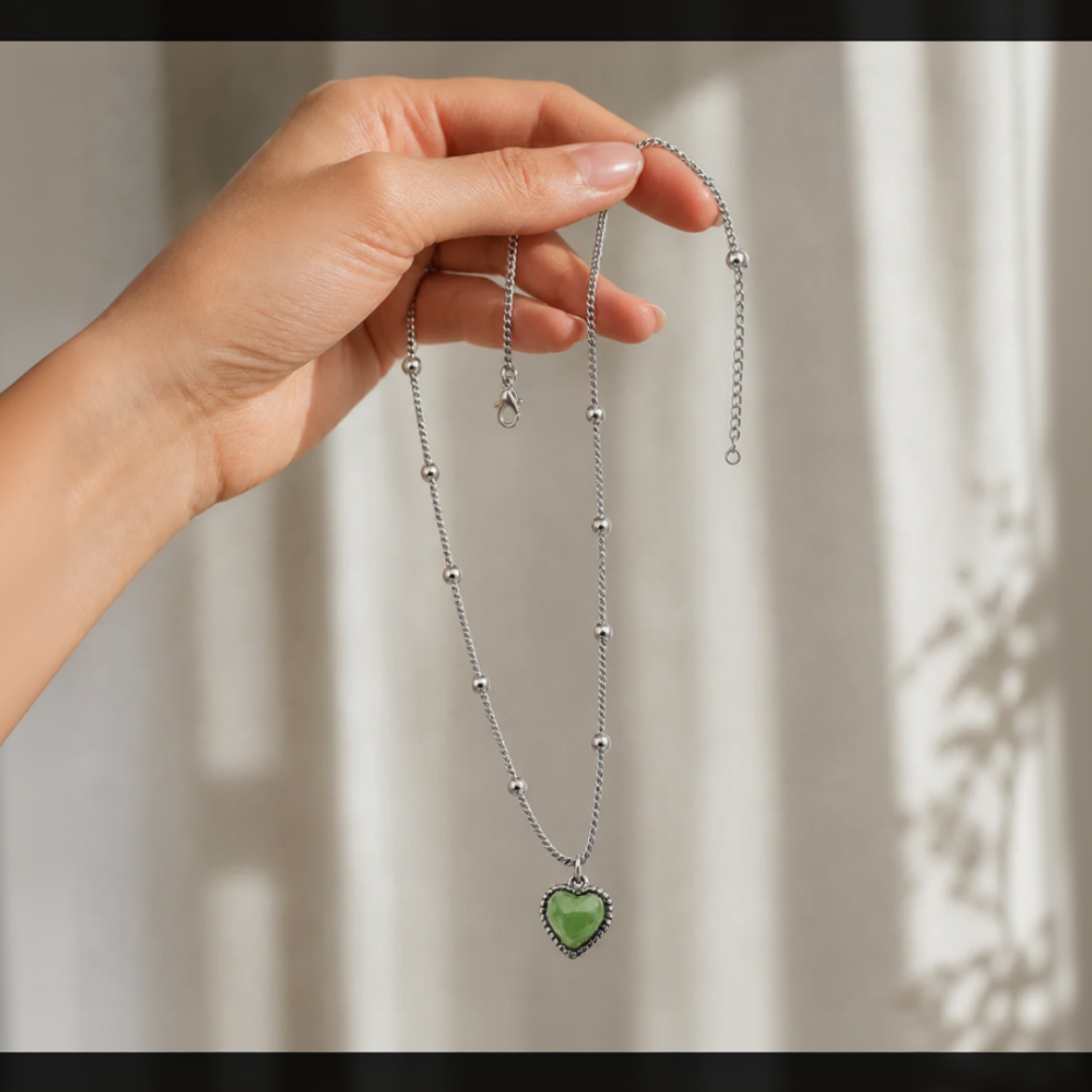 Hand holding a silver necklace with a green heart pendant against a neutral background